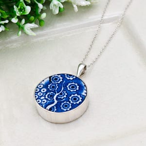 May include: A silver pendant necklace with a round, blue and white floral design. The pendant has a silver bezel and a delicate silver chain. The floral pattern includes stylized flowers and a tree-like motif. The necklace is displayed on a white surface.
