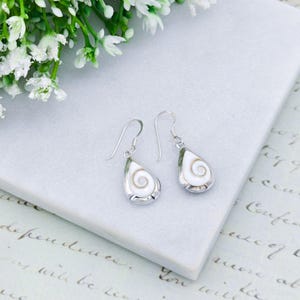 May include: Silver drop earrings with a teardrop design. Each earring has a white, spiral-patterned centre within a silver border. The earrings are displayed on a white surface, with white flowers in the background.