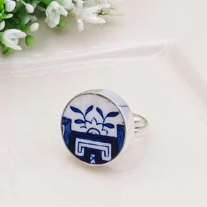 May include: A round silver ring with a blue and white floral design. The ring features a circular ceramic inlay with a detailed blue floral pattern against a white background. The ring band is silver.