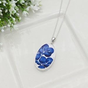 May include: Silver-toned necklace with an oval pendant featuring a blue floral design on a white background. The pendant is suspended from a delicate chain. The necklace is displayed on a white surface with white flowers in the background.