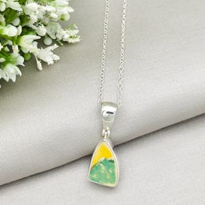 May include: A silver pendant necklace featuring a triangular stone with yellow and green hues. The stone is set in a silver bezel and hangs from a delicate silver chain. The necklace is displayed on a light gray surface.