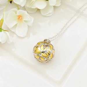 May include: A silver necklace featuring a gold and silver pendant. The pendant is a sphere with a floral pattern. The necklace is displayed on a white surface, with white flowers in the background.