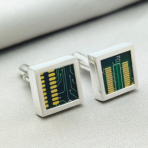 May include: A pair of silver cufflinks with a square design. Each cufflink features a green circuit board with yellow accents. The text "U2" is visible on one of the cufflinks.