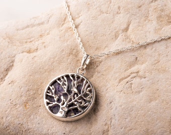 May include: A silver necklace with a round pendant featuring a tree design. The pendant has a dark purple background and a detailed, openwork tree silhouette. The necklace is displayed on a light-colored, textured stone.