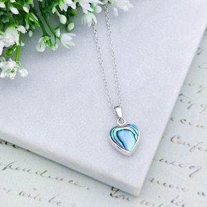 May include: A silver necklace with a heart-shaped pendant. The pendant features a blue and green iridescent abalone shell design. The necklace is displayed on a white surface with white flowers in the background.