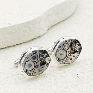 May include: Silver cufflinks displaying the internal workings of a watch. The oval-shaped cufflinks reveal gears, springs, and small jewels. The text "BULOVA WATCH CO" is visible on one. The background is a textured white surface.