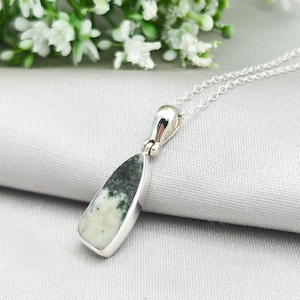 May include: A silver pendant necklace featuring a teardrop-shaped stone with a white and green pattern. The pendant is set in a silver bezel and hangs from a delicate silver chain. The necklace is displayed on a light gray surface.