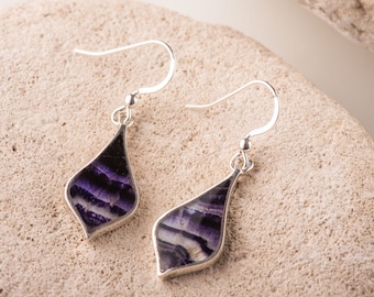 May include: A pair of silver dangle earrings with a teardrop shape. Each earring features a polished, bezel-set stone with a layered pattern of purple, white, and black. The earrings are displayed on a light-colored, textured surface.