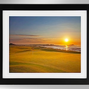 May include: A scenic view of a golf course with a green putting green in the foreground. The sun is setting over the ocean in the background, casting a golden glow over the landscape.