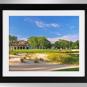 May include: A white building with a brown roof sits on a golf course with green grass and sand. The building is surrounded by trees and a blue sky with white clouds.