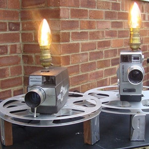 Könnte beinhalten: Zwei umgebaute Vintage-Filmkameras als Tischlampen mit Glühbirnen und Filmrollen als Sockel. Die Kameras sind silber und schwarz mit einem Retro-Design.