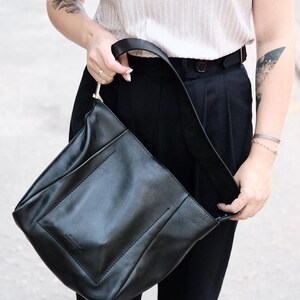 Puede incluir: Bolso de hombro de cuero negro con un dise&ntilde;o rectangular y una correa larga. El bolso tiene un estilo minimalista y un sutil detalle en relieve en la parte delantera. El bolso es sostenido por una persona que lleva una camisa clara y pantalones negros.