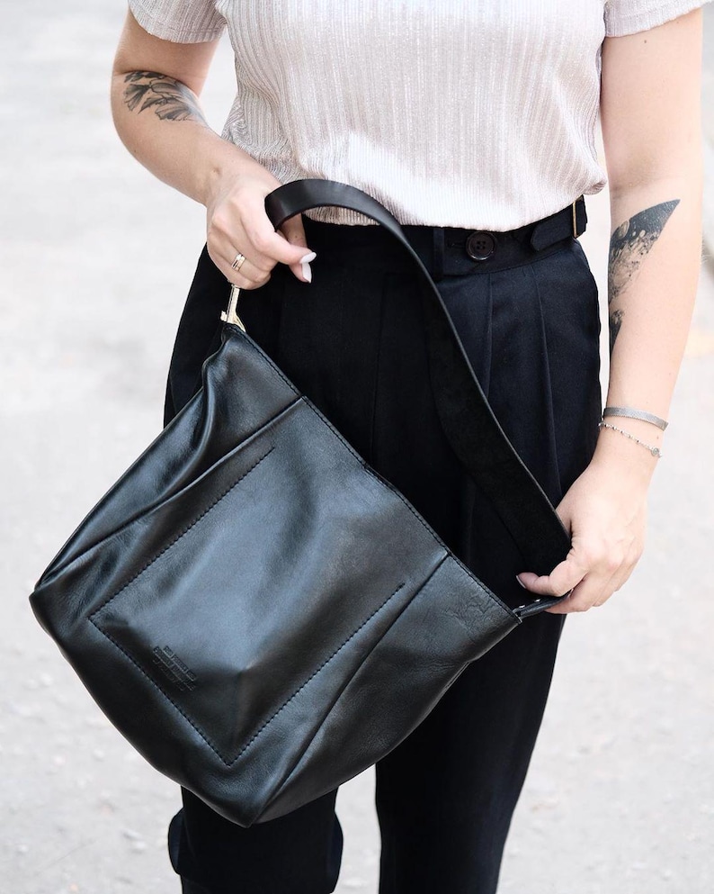 Puede incluir: Bolso de hombro de cuero negro con un dise&ntilde;o rectangular y una correa larga. El bolso tiene un estilo minimalista y un sutil detalle en relieve en la parte delantera. El bolso es sostenido por una persona que lleva una camisa clara y pantalones negros.