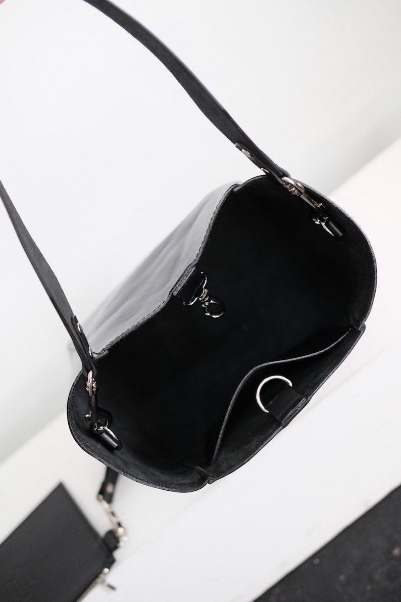 Puede incluir: Bolso tipo cubo de cuero negro con una correa larga para el hombro y una bolsa m&aacute;s peque&ntilde;a adjunta. El bolso tiene herrajes plateados y un dise&ntilde;o simple y minimalista. El interior tiene un peque&ntilde;o bolsillo.