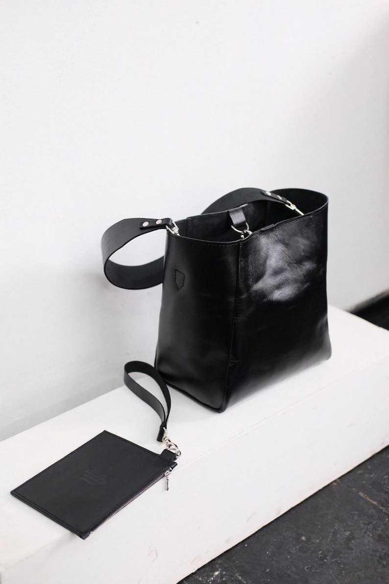 Puede incluir: Bolso tipo cubo de cuero negro con una peque&ntilde;a bolsa a juego. El bolso tiene una correa ancha para el hombro y un asa m&aacute;s peque&ntilde;a. La bolsa tiene cierre de cremallera y una correa de mu&ntilde;eca. El bolso y la bolsa est&aacute;n sobre una superficie blanca.