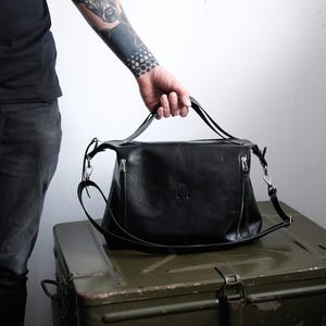 Pode incluir: Bolsa de ombro de couro preta com uma alça superior e uma longa alça ajustável. A bolsa tem formato retangular com fecho de zíper e um logotipo sutil em relevo na frente. A bolsa é segurada por uma pessoa com uma tatuagem na manga.