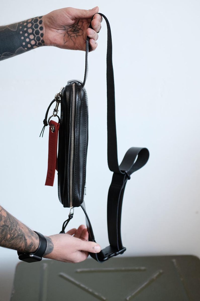 Puede incluir: Un bolso bandolera de cuero negro con cierre de cremallera y una etiqueta de cuero roja. El bolso tiene una correa ajustable con hebilla.