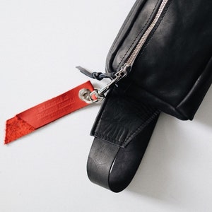 Puede incluir: Ri&ntilde;onera de cuero negro con cremallera plateada y una etiqueta de cuero roja que dice "The Original Handmade By" en letras blancas.