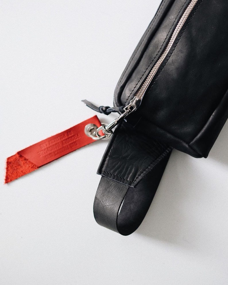 Puede incluir: Ri&ntilde;onera de cuero negro con cremallera plateada y una etiqueta de cuero roja que dice "The Original Handmade By" en letras blancas.