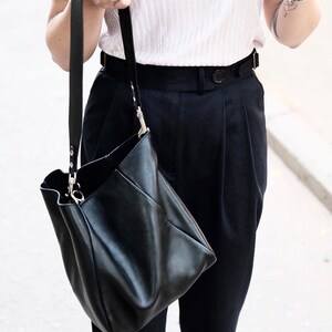Puede incluir: Un bolso de mano de cuero negro con una correa larga. El bolso tiene un dise&ntilde;o suave y holgado y herrajes plateados. La persona lleva una blusa de color claro y pantalones negros. El bolso es sostenido por una persona.