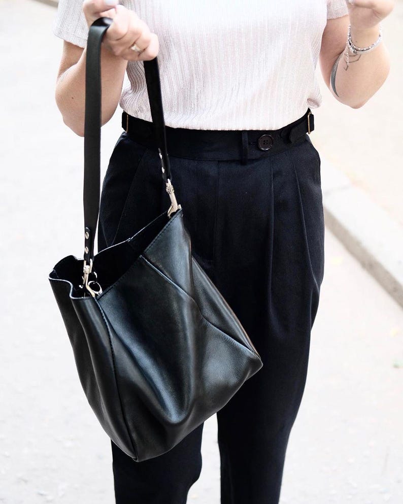 Puede incluir: Un bolso de mano de cuero negro con una correa larga. El bolso tiene un dise&ntilde;o suave y holgado y herrajes plateados. La persona lleva una blusa de color claro y pantalones negros. El bolso es sostenido por una persona.