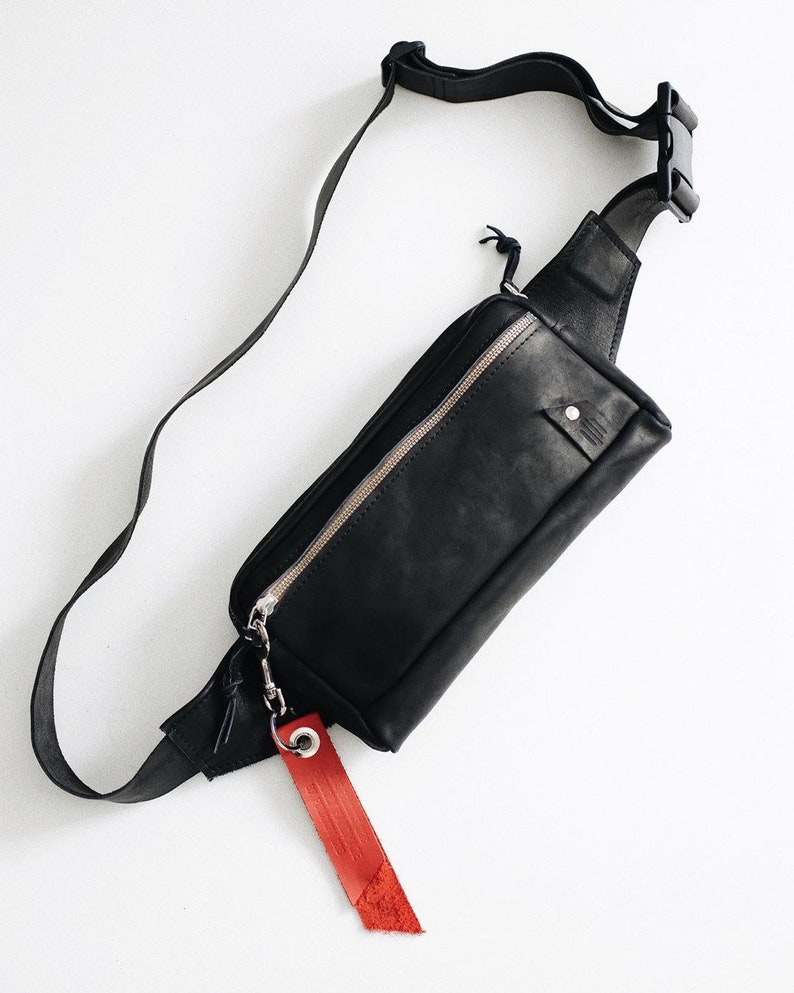 Puede incluir: Ri&ntilde;onera de cuero negro con cremallera plateada y etiqueta de cuero roja. La bolsa tiene una correa larga ajustable.