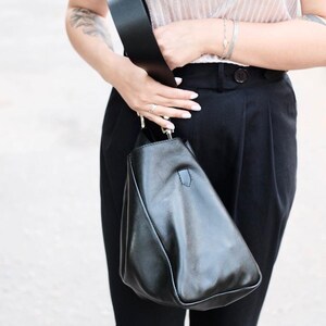 Puede incluir: Un bolso de hombro de cuero negro con una correa ancha. El bolso tiene una forma triangular &uacute;nica. La persona que lo sostiene lleva una parte superior de color claro y pantalones negros. La correa del bolso es gruesa y parece estar hecha del mismo material que el bolso.