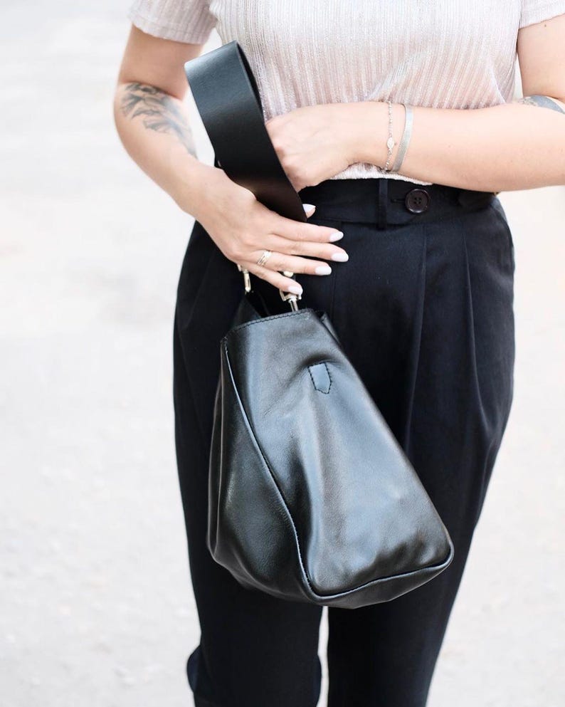 Puede incluir: Un bolso de hombro de cuero negro con una correa ancha. El bolso tiene una forma triangular &uacute;nica. La persona que lo sostiene lleva una parte superior de color claro y pantalones negros. La correa del bolso es gruesa y parece estar hecha del mismo material que el bolso.