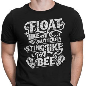 Könnte beinhalten: Schwarzes T-Shirt mit einem weißen Grafikdruck, auf dem "Float like a butterfly, sting like a bee" steht. Die Grafik enthält auch zwei Boxhandschuhe.