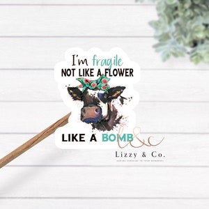 Puede incluir: Una pegatina con una ilustración de acuarela de una vaca con una diadema floral. El texto dice "Soy frágil, no como una flor, como una bomba" y "Lizzy & Co. Añadiendo sol a tus recuerdos."