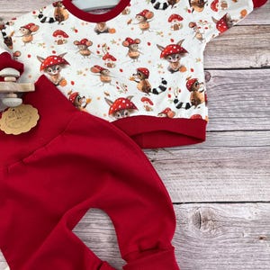 Könnte beinhalten: Ein zweiteiliges Baby-Outfit mit einem weißen Oberteil mit einem skurrilen Pilz- und Waldtierdruck, akzentuiert mit rotem Besatz. Die passende rote Hose hat eine bequeme, lockere Passform. Das Outfit wird auf einer Holzoberfläche präsentiert.