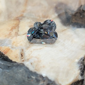 May include: A faceted, gray gemstone with a unique, cloud-like shape. The gem displays internal rainbow-colored flashes of light. It is set against a natural, light-colored stone surface.