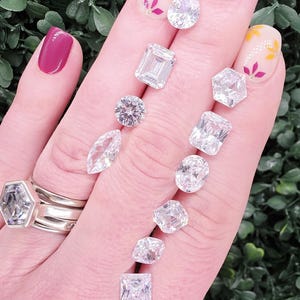 May include: A collection of ten loose, clear, faceted gemstones in various shapes including pear, emerald, round, marquise, oval, hexagon, and square cuts, displayed alongside a silver ring with a hexagonal stone.