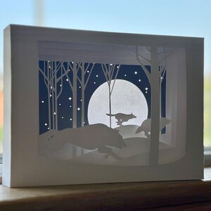 K&ouml;nnte beinhalten: Ein wei&szlig;es Papierkunstwerk, das W&ouml;lfe zeigt, die durch einen Wald unter einem Vollmond laufen. Die Szene ist geschichtet, um Tiefe zu erzeugen, mit Silhouetten von B&auml;umen und Sternen am Nachthimmel. Das Kunstwerk wird in einem wei&szlig;en Rahmen pr&auml;sentiert.