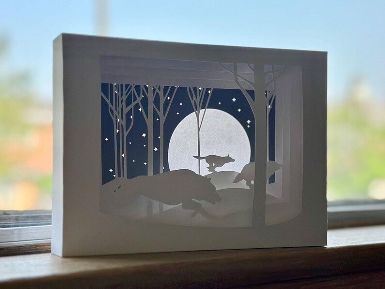 K&ouml;nnte beinhalten: Ein wei&szlig;es Papierkunstwerk, das W&ouml;lfe zeigt, die durch einen Wald unter einem Vollmond laufen. Die Szene ist geschichtet, um Tiefe zu erzeugen, mit Silhouetten von B&auml;umen und Sternen am Nachthimmel. Das Kunstwerk wird in einem wei&szlig;en Rahmen pr&auml;sentiert.