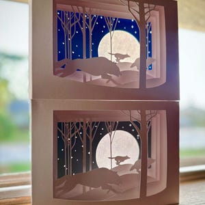 K&ouml;nnte beinhalten: Zwei geschichtete Papierkunstwerke, die eine n&auml;chtliche Waldszene darstellen. Silhouetten von W&ouml;lfen laufen &uuml;ber eine schneebedeckte Landschaft unter einem Vollmond und Sternenhimmel. Das Kunstwerk ist in einer hellrosa Box gerahmt.