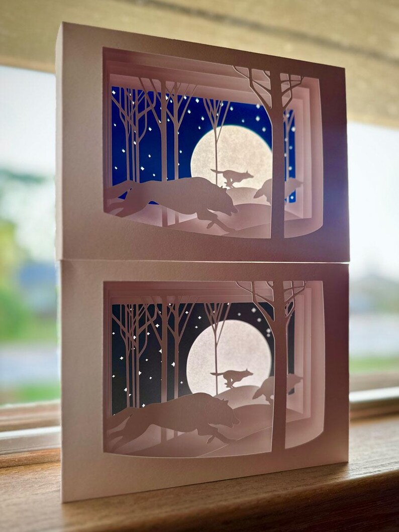 K&ouml;nnte beinhalten: Zwei geschichtete Papierkunstwerke, die eine n&auml;chtliche Waldszene darstellen. Silhouetten von W&ouml;lfen laufen &uuml;ber eine schneebedeckte Landschaft unter einem Vollmond und Sternenhimmel. Das Kunstwerk ist in einer hellrosa Box gerahmt.