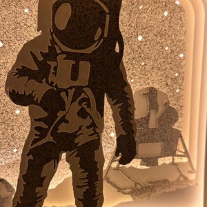 May include: A black silhouette of an astronaut standing on the moon with a lunar module in the background. The astronaut is wearing a spacesuit with a helmet and is holding a tool in one hand. The moon is depicted as a white surface with a starry sky.
