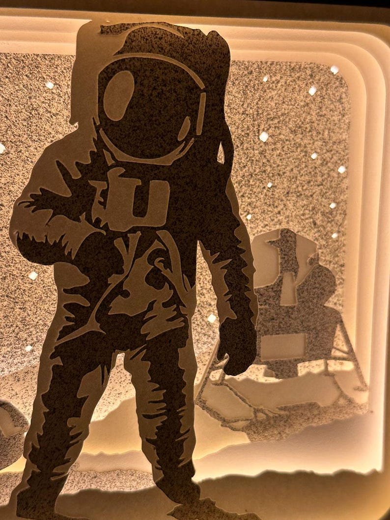 May include: A black silhouette of an astronaut standing on the moon with a lunar module in the background. The astronaut is wearing a spacesuit with a helmet and is holding a tool in one hand. The moon is depicted as a white surface with a starry sky.