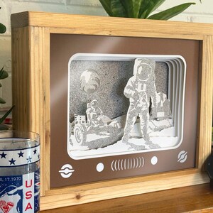May include: A wooden frame with a brown background and a white paper cutout of an astronaut on the moon. The astronaut is standing next to a lunar rover. The frame has a vintage television screen design with a white border and a black and white image of the moon landing.