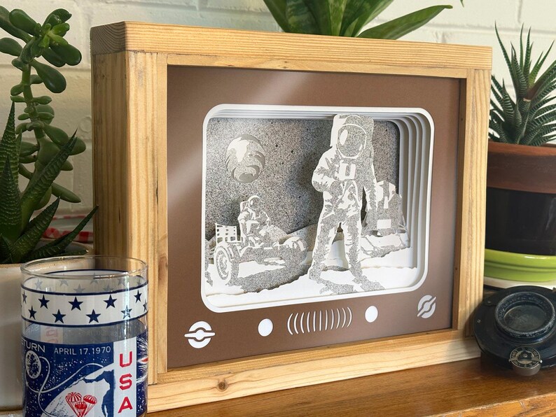 May include: A wooden frame with a brown background and a white paper cutout of an astronaut on the moon. The astronaut is standing next to a lunar rover. The frame has a vintage television screen design with a white border and a black and white image of the moon landing.