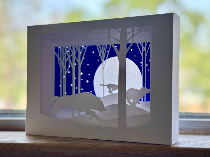 K&ouml;nnte beinhalten: Ein wei&szlig;es Papierkunstwerk, das eine n&auml;chtliche Waldszene darstellt. Silhouetten von laufenden Tieren befinden sich im Vordergrund, mit B&auml;umen, einem Vollmond und Sternen vor einem dunkelblauen Hintergrund. Das Kunstwerk wird in einem wei&szlig;en Rahmen pr&auml;sentiert.