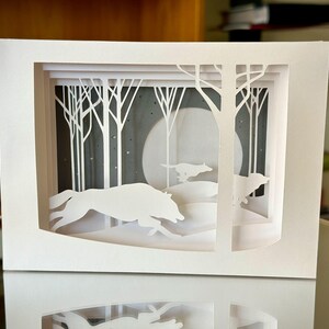 K&ouml;nnte beinhalten: Ein wei&szlig;es Papierkunstwerk, das eine Winterwaldszene darstellt. Silhouetten von W&ouml;lfen rennen durch den Schnee, mit B&auml;umen und einem Vollmond im Hintergrund. Das Kunstwerk ist geschichtet, um Tiefe zu erzeugen.