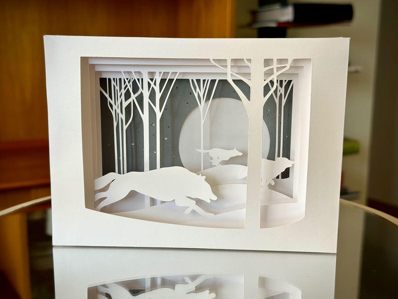 K&ouml;nnte beinhalten: Ein wei&szlig;es Papierkunstwerk, das eine Winterwaldszene darstellt. Silhouetten von W&ouml;lfen rennen durch den Schnee, mit B&auml;umen und einem Vollmond im Hintergrund. Das Kunstwerk ist geschichtet, um Tiefe zu erzeugen.