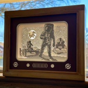 May include: A wooden frame with a red interior, depicting a 3D paper cut-out scene of an astronaut on the moon. The astronaut is wearing a spacesuit and is standing next to a lunar module. The scene is set against a starry background.