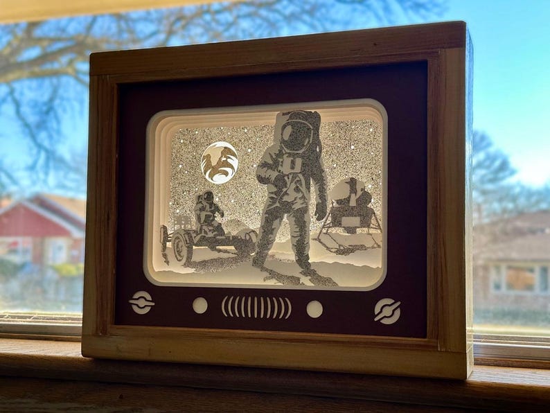 May include: A wooden frame with a red interior, depicting a 3D paper cut-out scene of an astronaut on the moon. The astronaut is wearing a spacesuit and is standing next to a lunar module. The scene is set against a starry background.