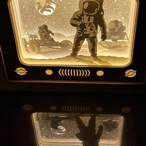 May include: A white wooden frame with a lit-up scene of an astronaut standing on the moon with a lunar rover. The scene is reflected in the surface below the frame.