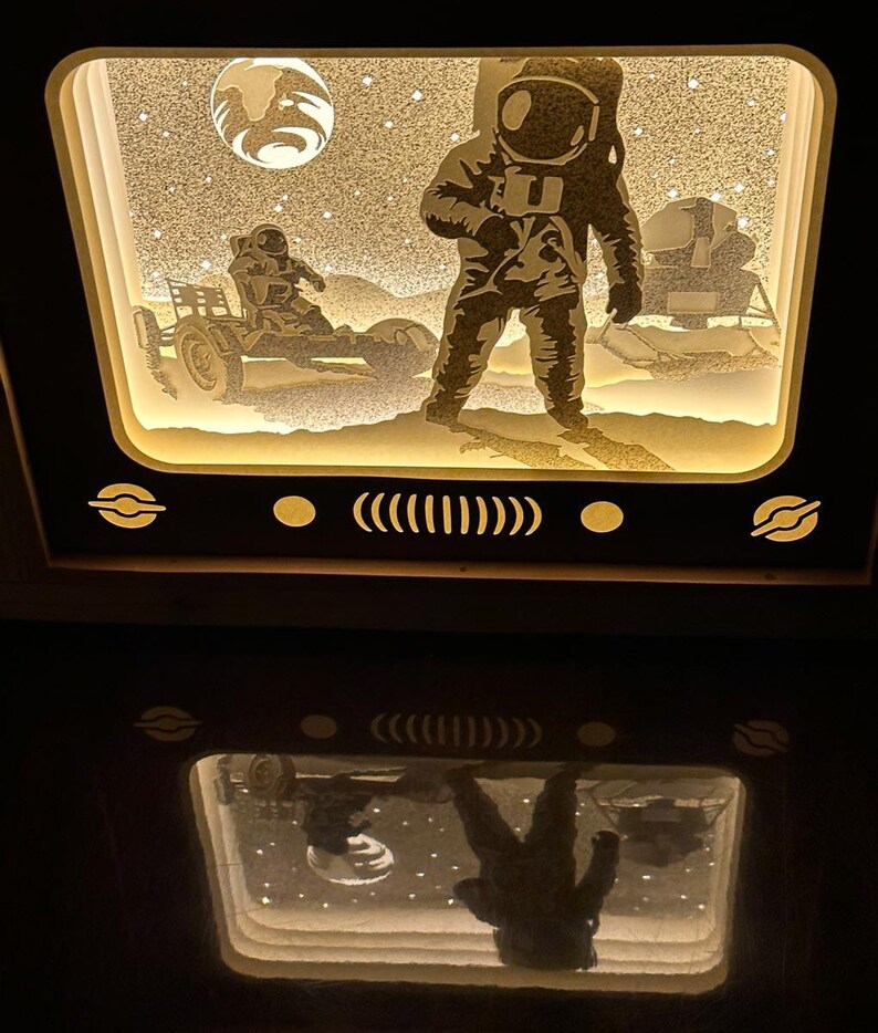 May include: A white wooden frame with a lit-up scene of an astronaut standing on the moon with a lunar rover. The scene is reflected in the surface below the frame.