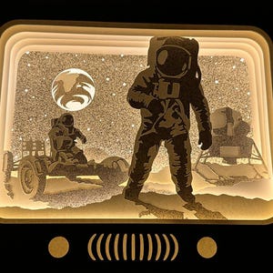 May include: A 3D layered paper art piece depicting an astronaut standing on the moon's surface. The astronaut is wearing a white spacesuit with a helmet. The moon's surface is textured with craters and a small lunar rover is visible in the background. The scene is illuminated with a warm white light.
