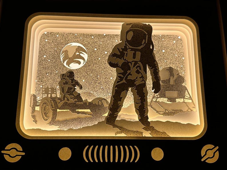May include: A 3D layered paper art piece depicting an astronaut standing on the moon's surface. The astronaut is wearing a white spacesuit with a helmet. The moon's surface is textured with craters and a small lunar rover is visible in the background. The scene is illuminated with a warm white light.
